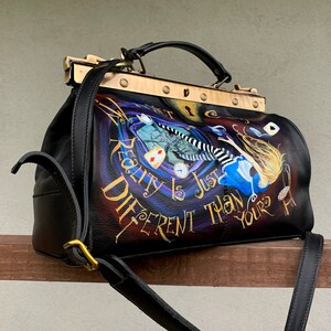 May include: Black leather doctor bag with a gold metal clasp and strap. The bag features a painted design of Alice falling down a rabbit hole with the text "Reality is just different than yours".