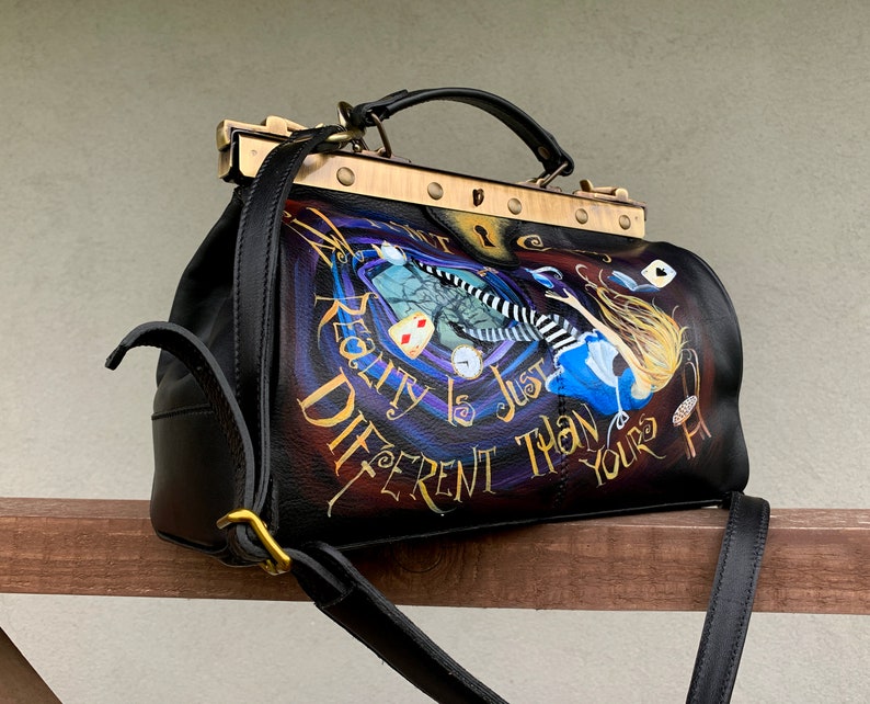 May include: Black leather doctor bag with a gold metal clasp and strap. The bag features a painted design of Alice falling down a rabbit hole with the text "Reality is just different than yours".