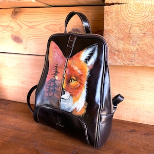 Puede incluir: Una mochila de cuero marrón oscuro con un diseño pintado a mano en el panel frontal. La obra muestra una composición dividida con un paisaje forestal al atardecer a la izquierda y un retrato detallado de un zorro rojo en el lado derecho.