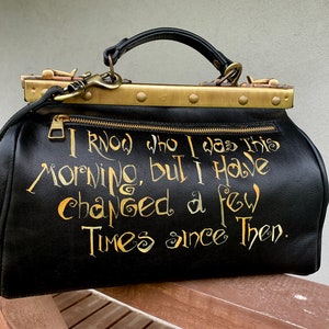 May include: Black leather doctor bag with gold hardware and a gold inscription that reads "I know who I was this morning, but I have changed a few times since then."