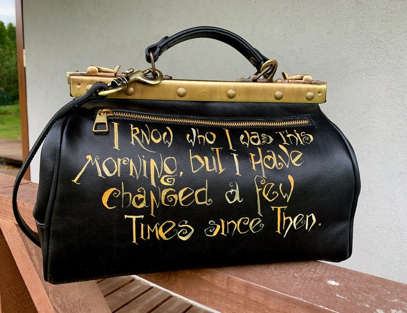 May include: Black leather doctor bag with gold hardware and a gold inscription that reads "I know who I was this morning, but I have changed a few times since then."