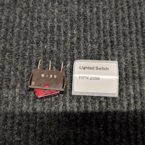 May include: A brown rocker switch with a red translucent actuator and three metal contact pins. The switch body features the printed text 0 1 3 8. Beside it is a white rectangular label with the text Lighted Switch and MPN 2556.