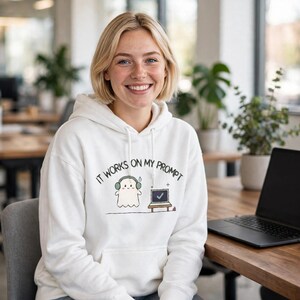Puede incluir: Una sudadera con capucha blanca que presenta un gráfico de un pequeño fantasma con auriculares verdes junto a un portátil que muestra una marca de verificación verde. El texto superior dice IT WORKS ON MY PROMPT en letras negras.