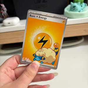 May include: A rectangular Pokémon trading card featuring a bright yellow background with a large black lightning bolt symbol. The text reads Basic Energy at the top. A blue and cream-coloured creature with a fluffy coat is illustrated at the bottom.