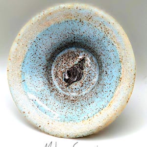 May include: A round ceramic bowl with a light blue glazed interior featuring brown speckles and a dark, embossed seashell design at the center. The rim is textured with a sandy, beige finish. The text Malama Ceramics is printed below.
