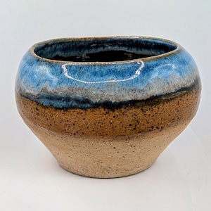 May include: A round ceramic bowl with a wide opening, featuring a two-tone glaze. The upper portion is a glossy, mottled blue that transitions into a dark band, while the lower section is a textured, speckled tan and brown clay finish.