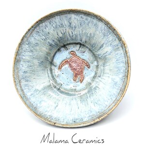 May include: A round ceramic bowl with a mottled light blue and white glaze. The center features a raised, copper-toned sea turtle relief. The rim is a natural tan color. Below the bowl, the text Malama Ceramics is printed in black.
