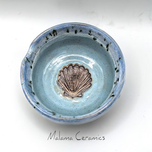 May include: A round ceramic bowl with a light blue speckled glaze and a dark brown embossed seashell design at the center. The rim features a darker blue tone with black speckles. The text Malama Ceramics is printed below.