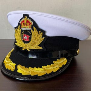 Könnte beinhalten: Eine Schirmmütze mit weißem Oberteil, schwarzem Band und schwarzem Schirm. Vorne befindet sich ein goldenes Stickemblem mit einer Krone über einer roten Flagge mit weißem Symbol. Der schwarze Schirm ist mit goldenem Blattwerk bestickt.