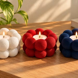 Op de afbeelding: Drie ronde kaarsenhouders op een rij, elk met een textuur van kleine bolletjes. De houders zijn wit, rood en donkerblauw en bevatten elk een klein, brandend waxinelichtje in het midden van het bolvormige ontwerp.