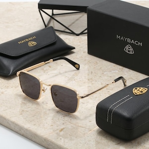 May include: Gold-tone metal sunglasses with dark grey lenses and black temple tips. The frames feature a square shape. Included are a black leather-like pouch and a black rectangular box, both branded with the Maybach logo and text in gold.