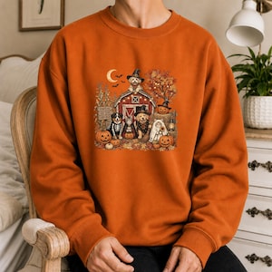 Pode incluir: Uma camisola laranja de gola redonda com uma impressão gráfica de um celeiro vermelho rodeado por folhagem de outono, abóboras e vários cães e gatos vestidos com fatos de Halloween, incluindo chapéus de bruxa e um lençol de fantasma, sob uma lua crescente.
