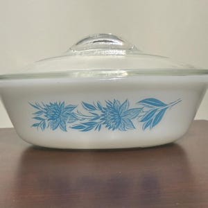 Puede incluir: Una fuente de horno ovalada de color blanco con una tapa de cristal transparente. El exterior de la fuente presenta un patrón floral azul repetitivo que consiste en flores y hojas estilizadas dispuestas en una banda horizontal.