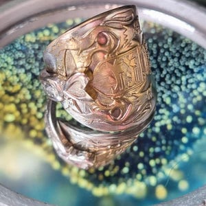 May include: A silver-toned metal spoon ring featuring an intricate embossed design with a central shield motif, floral patterns, and a heart-shaped detail. The band is formed from the handle of a vintage spoon, showing metallic textures.