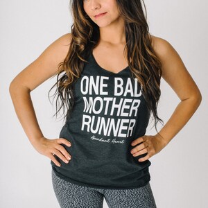 May include: A woman wearing a black tank top with the text "ONE BAD MOTHER RUNNER" and the words "Abundant Heart" underneath. She is also wearing gray and black patterned leggings.