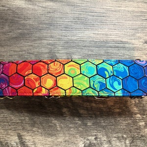 K&ouml;nnte beinhalten: Ein Hundehalsband mit einem bunten Regenbogen-Wabenmuster. Das Halsband hat ein lebendiges Design mit Farbt&ouml;nen von Rot, Orange, Gelb, Gr&uuml;n, Blau und Lila. Es hat eine Metallschnalle und einen D-Ring zur Befestigung der Leine.