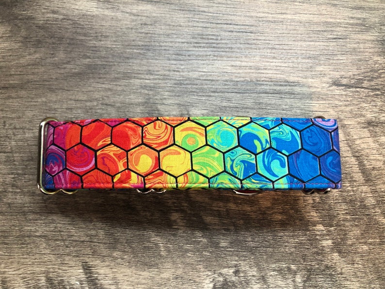 K&ouml;nnte beinhalten: Ein Hundehalsband mit einem bunten Regenbogen-Wabenmuster. Das Halsband hat ein lebendiges Design mit Farbt&ouml;nen von Rot, Orange, Gelb, Gr&uuml;n, Blau und Lila. Es hat eine Metallschnalle und einen D-Ring zur Befestigung der Leine.