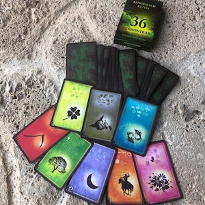 May include: A deck of 36 Lenormand cards with a box labeled 36 PERSONĪBAS. The cards feature colorful illustrations including a clover, mountain, fish, tree, moon, rider, and flowers, with dark green backs and black borders on a textured surface.