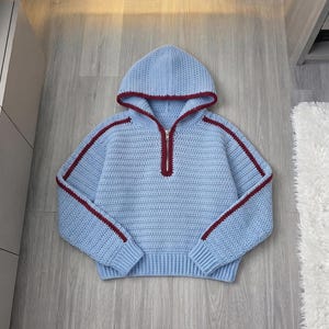 May include: A light blue crochet hoodie featuring a half-zip front closure with a silver zipper. The garment is accented with dark red trim along the hood edge, the zipper placket, and a single vertical stripe running down each sleeve.