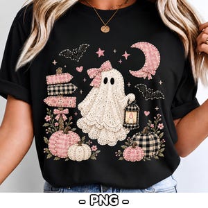 Può includere: Una t-shirt nera con la grafica di un fantasma in pizzo bianco che indossa un fiocco rosa e tiene in mano una lanterna. Il design include zucche a quadri rosa e neri, un cartello in legno, pipistrelli, una luna crescente e piccoli fiori rosa.