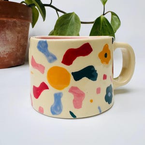 Puede incluir: Una taza de cerámica con una base de color crema que presenta un patrón disperso de formas abstractas en rojo, amarillo, azul claro, azul oscuro y rosa. La taza tiene un asa resistente en el lado derecho y un acabado suave y brillante.