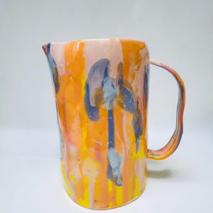 Könnte beinhalten: Ein Keramikkrug mit einer leuchtenden, mehrfarbigen Glasur mit vertikalen Tropfen und Wirbeln in leuchtendem Orange, Gelb, zartem Rosa und gedämpftem Blau. Das Gefäß hat einen abgerundeten Körper, einen geschwungenen Henkel und einen kleinen Ausguss.