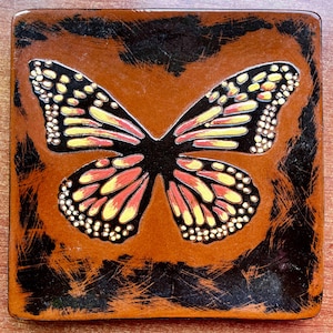Puede incluir: Un posavasos cuadrado con una ilustración central de una mariposa monarca con alas negras, naranjas y amarillas. El fondo es una mezcla texturizada y desgastada de tonos marrón oscuro y negro, creando un aspecto rústico en la superficie.