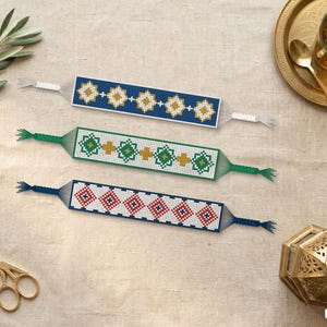 May include: Three rectangular woven bracelets featuring geometric diamond patterns. The top bracelet is blue with gold diamonds, the middle is white with green and gold diamonds, and the bottom is white with red and blue diamonds.