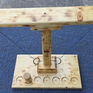 May include: A wooden ring toss game featuring a rectangular base with six circular indentations, a central vertical post, and a horizontal crossbar. Two metal rings are attached to the crossbar by thin strings, and one small wooden cylinder sits in a base slot.