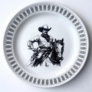 May include: White ceramic plate with a black and white illustration of a cowboy on a horse. The plate has a decorative gray border. The cowboy is wearing a hat and holding the reins. A decorative item.