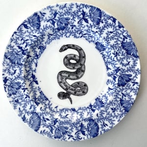 Peut inclure: Une assiette ronde en céramique présentant une illustration centrale en noir et blanc d'un serpent enroulé. Le bord est orné d'un motif floral détaillé bleu et blanc composé de vignes et de fleurs sur un fond blanc.