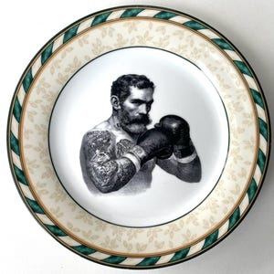 Może przedstawiać: Okrągły talerz ceramiczny z czarno-białą ilustracją brodatego boksera z tatuażami na ramionach i klatce piersiowej, ubranego w rękawice bokserskie. Brzeg jest kremowy z motywem liści oraz zielono-białym paskiem na samej krawędzi.