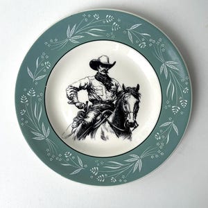 May include: A vintage decorative plate with a black and white illustration of a cowboy on horseback. The plate has a teal border with a white floral design. The centre of the plate is white.