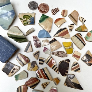 May include: A collection of antique pottery shards in various shapes and sizes, featuring colorful patterns and designs. The fragments include blue, green, red, and black accents, with two coins for scale. The pottery pieces are scattered on a white surface.
