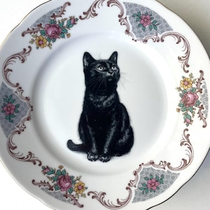 May include: A white ceramic plate featuring a detailed illustration of a black cat sitting in the center. The rim is decorated with a repeating pattern of pink roses, yellow flowers, and grey lattice accents with gold-toned edges.