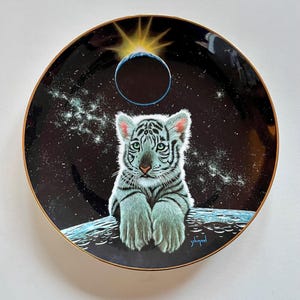 May include: A white porcelain plate with a gold rim features a white tiger cub sitting on a moon-like surface. The background is a black starry night with a blue planet and a sunburst.