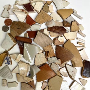 Op de afbeelding: Een verzameling gebroken aardewerkscherven in verschillende vormen en maten. De fragmenten vertonen een reeks kleuren, waaronder wit, bruin, beige en rood. Sommige stukken hebben zichtbare patronen en texturen, wat suggereert dat ze afkomstig zijn van borden, kopjes of andere keramische items.
