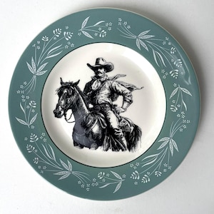 May include: A decorative plate with a teal rim and a central black and white image of a cowboy on horseback. The rim has a white floral pattern. The plate has a white centre.