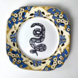 May include: Square decorative plate with a white center featuring a detailed black and gray snake illustration. The rim has a cream background with blue floral and gold accents. The plate has a gold trim.