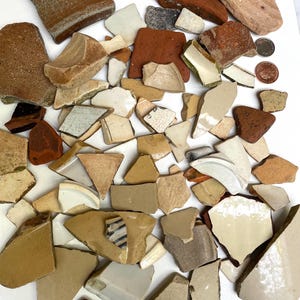 May include: A collection of broken pottery shards in various shapes and sizes. The fragments display a range of colors, including beige, brown, red, and white. Some pieces have visible patterns or textures, and a few coins are also present.