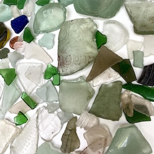 May include: A collection of sea glass shards in various shapes and sizes. The pieces are primarily shades of green, white, and clear, with a few blue and brown fragments. Some pieces have visible markings or text.
