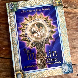 Könnte beinhalten: Ein Buch mit dem Titel "The Merlin Mystery" mit blauem und goldenem Einband. Das Cover zeigt ein Sonnenstrahl-Design mit einem silbernen Stab und dem Text "The Secret Lies Inside". Dekorative Eckakzente und keltische Knotenbordüren.