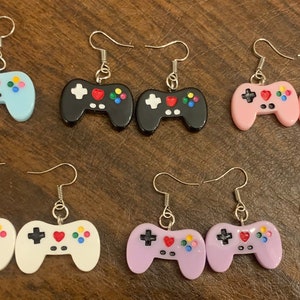 May include: Six pairs of colorful video game controller earrings. Each pair is a different color: light blue, black, pink, white, purple, and red.