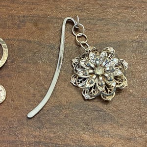 May include: Three metal bookmarks with decorative charms. One has a circle and crescent moon charm, another a flower, and the last an elephant. The bookmarks are silver-toned and rest on a brown surface.