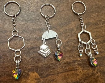 May include: Three silver keychains with different charms. The first keychain has a hexagonal charm with a heart-shaped crystal. The second keychain has a charm with a stack of books and a heart-shaped crystal. The third keychain has a hexagonal charm with a heart-shaped crystal and several smaller crystal charms.