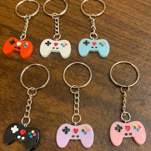 May include: Seven colorful plastic video game controller keychains. The keychains are red, white, light blue, black, lavender, and pink. Each keychain has a silver metal ring and chain.