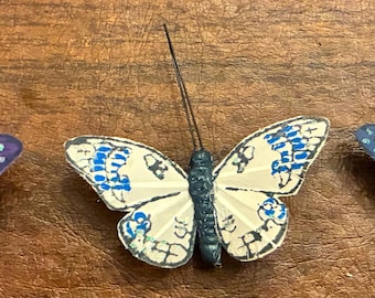 May include: Three decorative butterfly ornaments. One is purple with blue accents, the second is cream with blue details, and the third is blue with white spots. Each has a dark body and antennae.