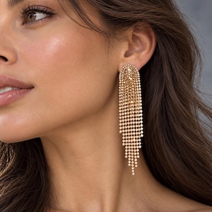 May include: A long, gold-toned dangle earring featuring a semi-circular top stud connected to multiple cascading strands of small, reflective metallic beads that create a tiered, fringe-like effect hanging down from the earlobe.