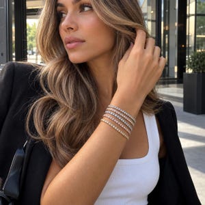 Puede incluir: Una pila de cinco pulseras de tenis en la muñeca, con una mezcla de engastes de metal en tono dorado, oro rosa y plateado. Cada pulsera presenta una hilera continua de pequeñas piedras transparentes de corte redondo que reflejan la luz.