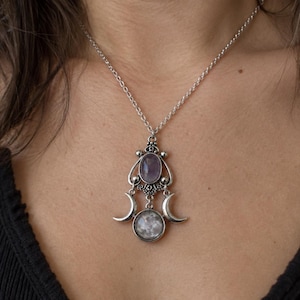 May include: A silver-toned necklace featuring a pendant with an oval purple stone at the top, a circular moon-patterned stone at the bottom, and two crescent moon accents on the sides, all suspended from a delicate silver-toned chain.
